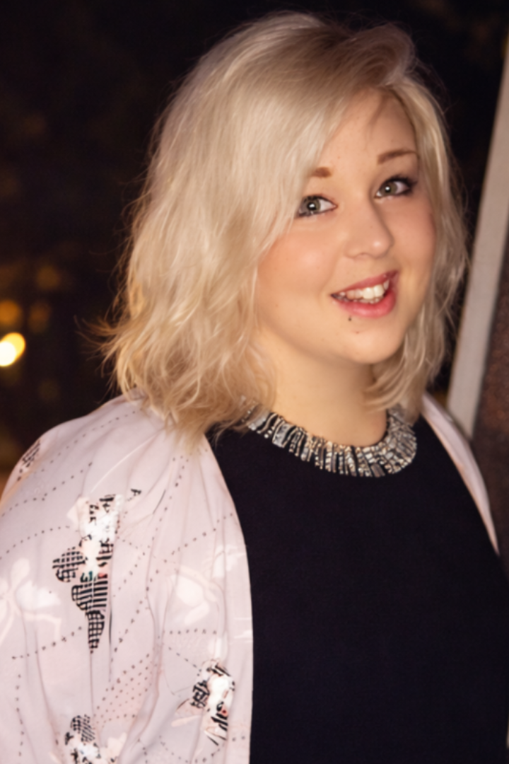 A woman is wearing a pink jacket and a black top, with a necklace. She has blonde hair and is smiling at the camera.