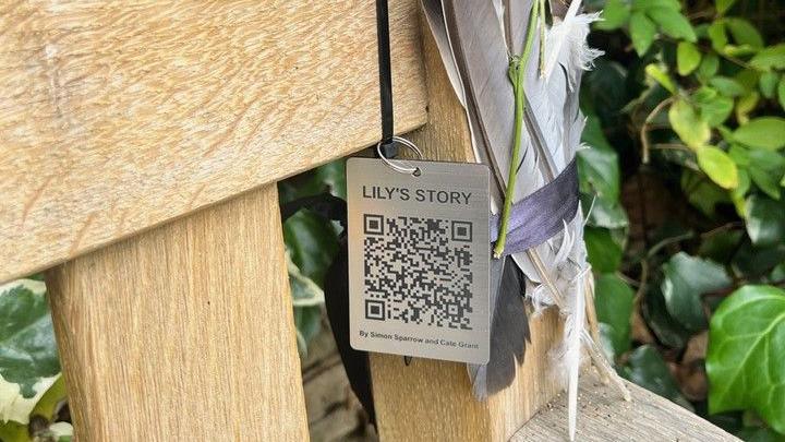 A grey, metal tag with a QR code engraved underneath the words Lily's Story. It is attached to the corner of a wooden bench. There are also feathers above the armrest.