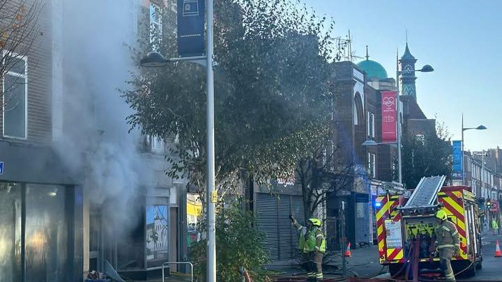 Fire crews and an engine on a street in Clacton-on-Sea. Smoke is coming from a building and fire officers are looking and pointing at the building, which is in street full of shops.