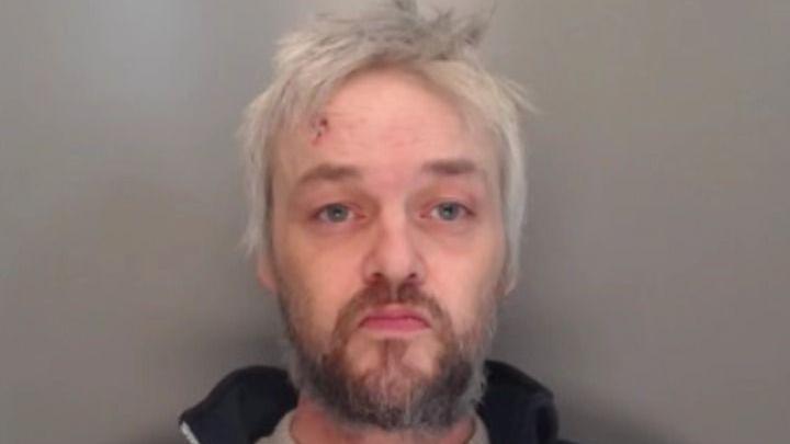 Mugshot of Tate. He has long messy straggly white hair and an untidy dark beard. He appears to have a cut on his forehead