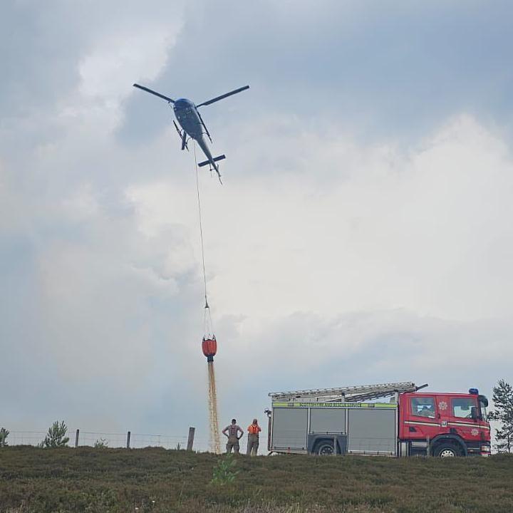 Two firefighters standing next to a fire engine watch as a helicopter with a bucket on the end of a long cable waterbombs a fire. The fire is not visible above the crest of a hill, but smoke can be seen rising from it.