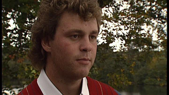 Darren Clarke stands in front of a tree, beside a lake. He is wearing a red rugby shirt with a white collar.