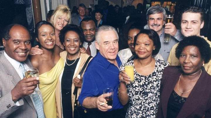 A group of people pictured with drinks inside a club, all of them smiling at the camera. Dr Stephenson is on the far left of the image, while Tony and Lalel Bullimore are in the centre wearing a bright blue shirt and a black and white flowery dress respectively. 