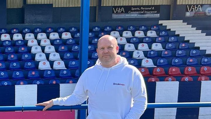 Dan Chandler with a hoodie and jeans stood in a football stadium