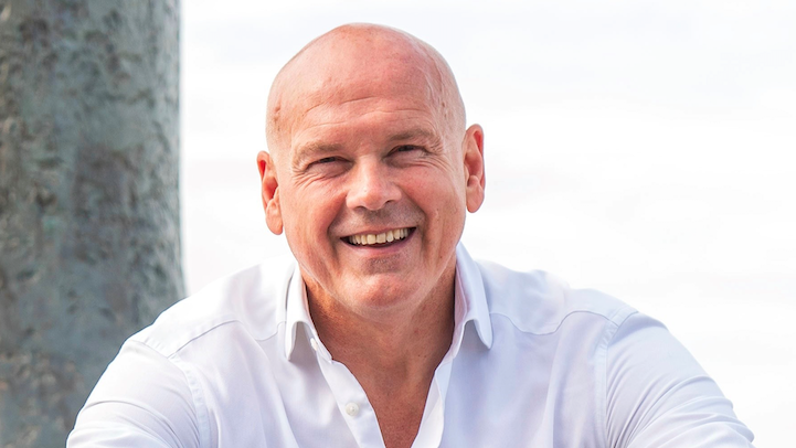 A bald man smiling, wearing a white collared shirt and white skies in the background.