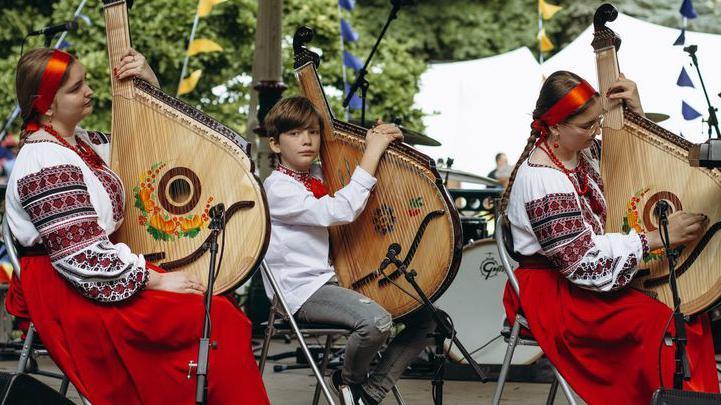 Three musicians at 2024's UkraineFest sat in a row in traditional dress of red and white preparing to play large stringed instrument.