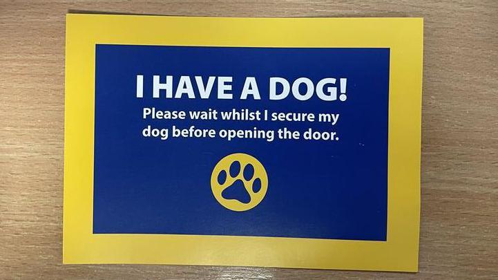 The blue and yellow card given to customers. It says "I have a dog, please wait whilst I secure my dog before opening the door". 
