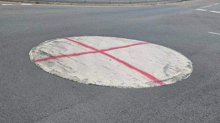 A white circular roundabout in the middle of a road, with a red cross painted on it, depicting the St George's Cross