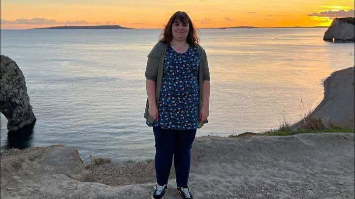 Shanice, who wears dark jeans, a long top and cardigan, smiles at the camera. She has short brown hair and a fringe, and is posing in front of a beautiful sunset view of the coast.
