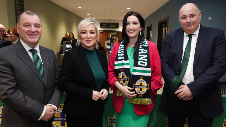 Patrick Nelson, a man with short, dark hair, wearing a grey suit and a green and navy IFA tie, stands smiling beside First Minister Michelle O'Neill who has blonde hair and is wearing a black suit over a dark green top, and Deputy First Minister Emma Little-Pengelly who has long, dark hair and is wearing a Northern Ireland football scarf over a green dress and red jacket. IFA President Conrad Kirkwood is standing beside Little-Pengelly wearing a navy suit and a dark green tie.