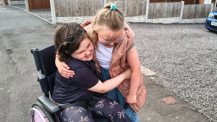 Sophia hugs Alice as she is sat in a wheelchair. Sophia has dark patterned leggings on and a black top. Alice has a beige body warmer on with a white T'shirt on.
