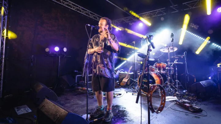 A man in a shirt standing on stage in front of a microphone and drum kit