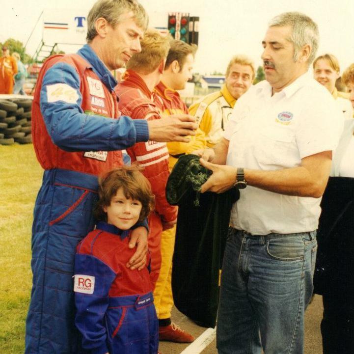 Abbie Eaton as a young girl dressed in a red and blue racing suit at a racing circuit. She is being held by her father, who is wearing a matching suit and holding a trophy presented by a man in a white shirt and blue jeans.