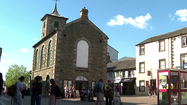A general view of Moot Hall in Keswick. It is in the middle of a pedestrian area and has an arched entrance with a sign for the tourist information office.
The brick building has a turret at the back, topped by a weather vane.