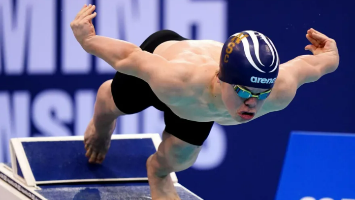 Bruce Dee wearing black swimming trunks, goggles and a swimming cap, dives into a pool 