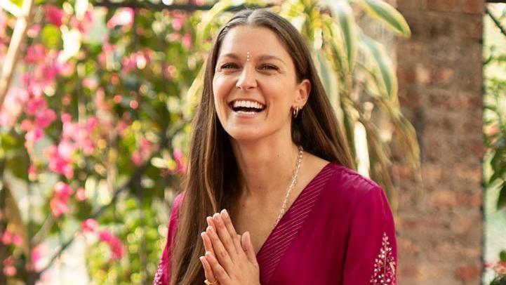 Charlotte Holmes smiles with her mouth open while sitting on a yoga mat, with her long brown hair over her shoulder and her hands pressed in a prayer position while she wears a purple dress