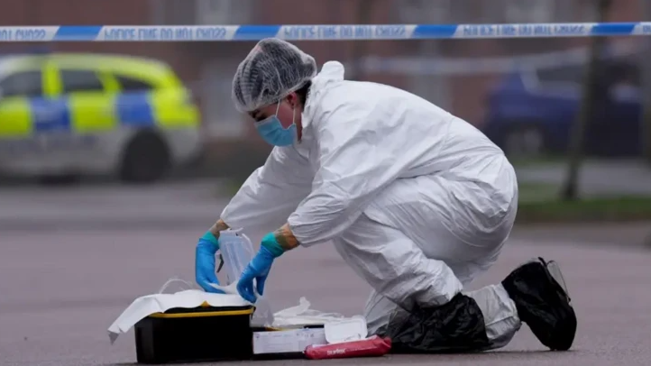 A forensics officer in a white suit and wearing blue gloves, a mask and a hairnet kneels in a road gathering evidence. There is police tape behind her and a police vehicle in the distance.