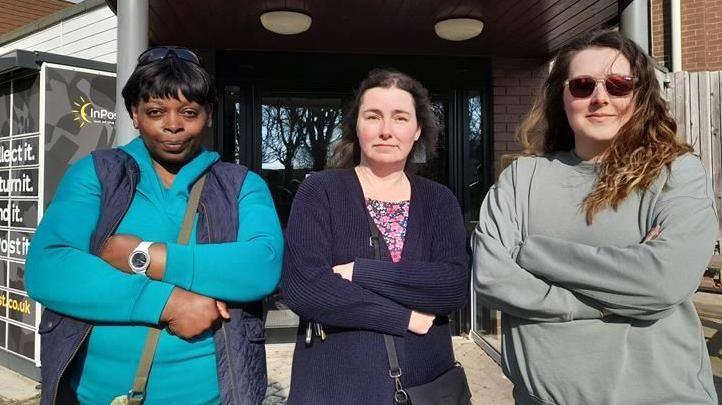 Three women stand outside the front door of a closed leisure and sports centre looking unhappy and disappointed. All have their arms folded and are looking directly at the camera.