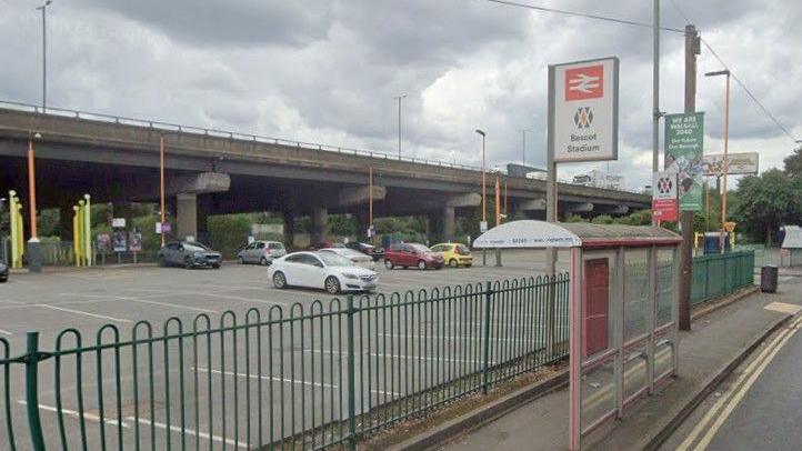 A car park next to a concrete bridge carrying a motorway. Several cars are parked inside behind green, metal railings. A sign next to the fence says "Bescot Stadium" and there is a bus stop in front of it.