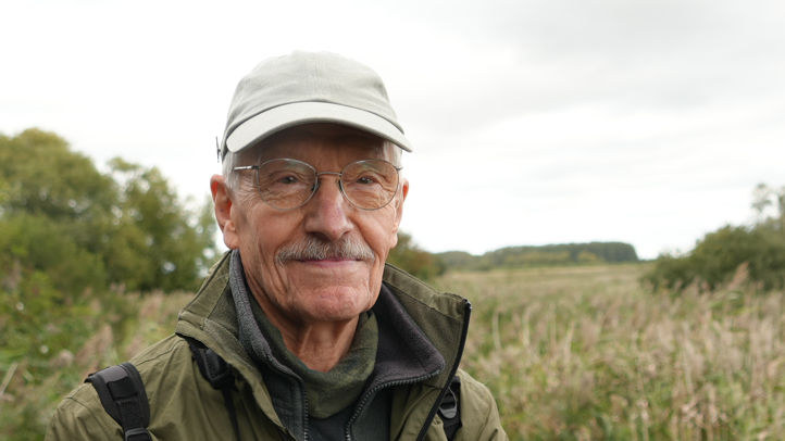 A man wearing a cap, glasses and a green jacket. He is smiling into the camera. There is grassland in the background.