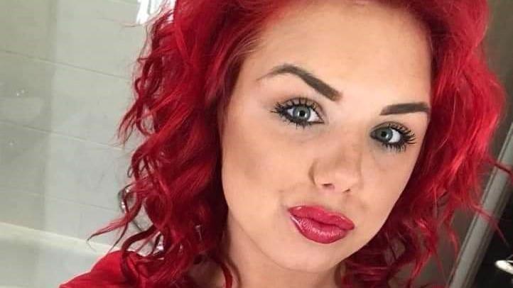 Selfie head shot of a pouting woman with bright dyed red hair. She is waring a red top and the image appears to have been taken in a bathroom.