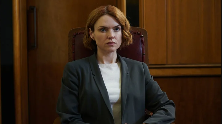 Erin with short curled ginger hair, wearing a grey suit and white t-shirt, sat on a red chair with a wooden wall behind her. She has a serious expression as she looks off to the side.