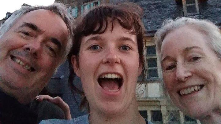 A close-up smiling selfie of a young woman alongside her grinning parents. All three are pictured from the neck up and are standing in front of Mont-Saint-Michel, a tidal island and mainland commune in Normandy, France. 