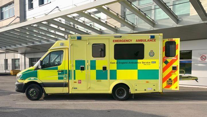 A yellow ambulance with green patches. The words "Emergency ambulance" are visible in red lettering at the top of the side of the ambulance. The trust's name is in black writing half way down the side. A rear door, with red stripes, is open. The ambulance is parked outside a hospital.
