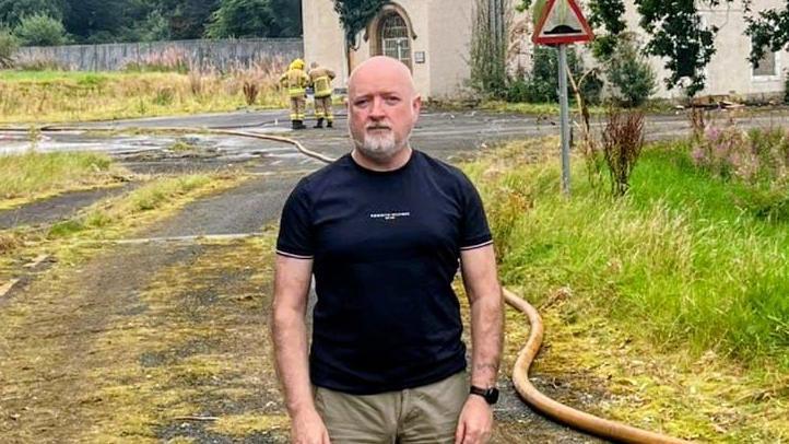 Shows a man with a bald head and goatee and black t-shirt standing in front of a building damaged by fire. A fire engine to the left and some firefighters in full kit. To the right there are hoses and a sign saying "Ramps".