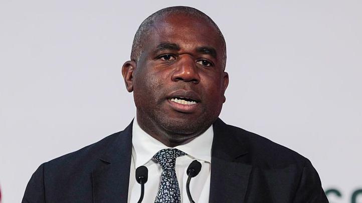 David Lammy speaks in front of a white background, wearing a white shirt, dark blazer and flowery tie.