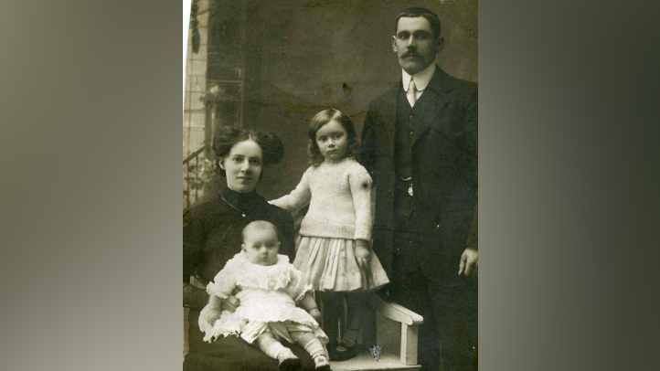 A black and white photo of Mary Schweermann holding her baby daughter Edna. Her older daughter Delia is standing on a chair next to them. She has her arm around her mother's shoulders. Their dad, Herbert Vince, is standing next to Delia. Mary is wearing a long black dress with high neck and long sleeves. Her black hair is styled in two high buns. Baby Edna is wearing white socks and a white dress. Delia is wearing a white jumper and a white skirt. Her curly hair reaches her shoulders. Herbert is wearing a black suit with a waistcoat, white shirt and a white tie. He has short black hair and a mustache.
