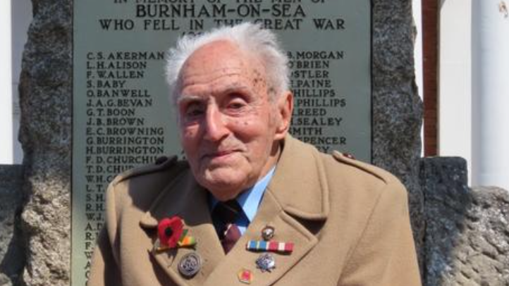 An elderly man wearing a brown coat with medals and a poppy on