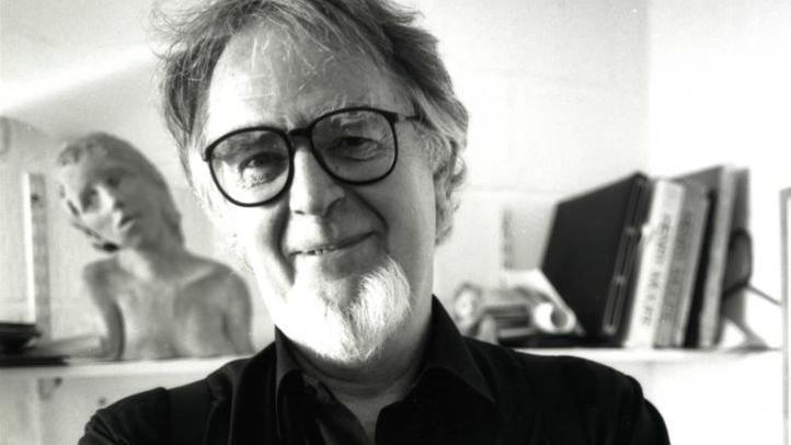A black and white photo of Bryan Kneale smiling. he has dark hair and a white beard, and is wearing black rimed glasses and a black shirt. There is a shelf behind him that has a sculpture of and some books on it.