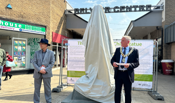 Two men in suits, the one on the left wearing civic chains of office, stand either side of a statue covered by a sheet.