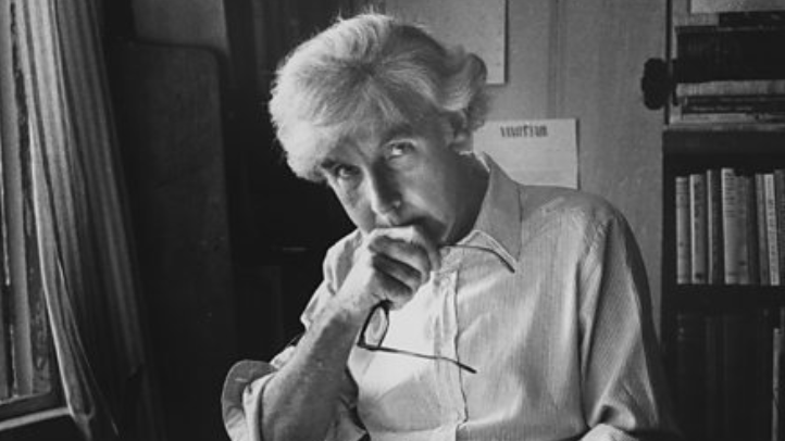 Ronald Blythe pictured in black and white. He is sitting a desk with books open. He is holding his glasses and looking into the camera. 