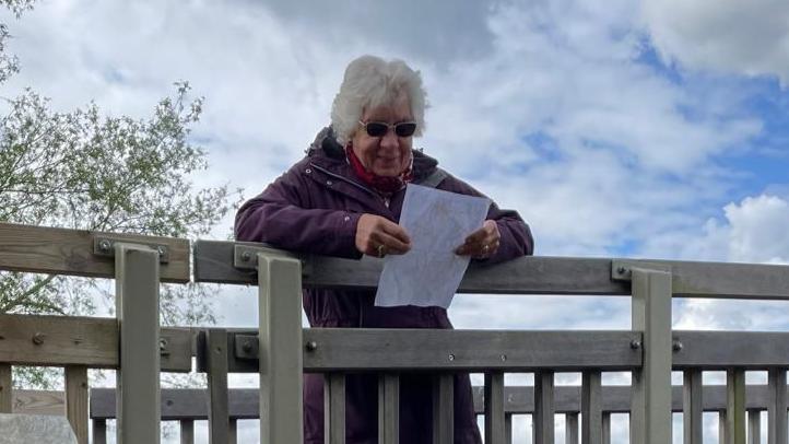 Mrs Savage rests over a bridge looking at a piece of paper. She has short grey hair and wears sunglasses.