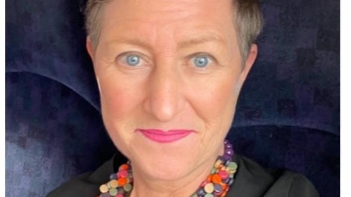 A woman with short hair sits smiling on a blue velvet chair. She wears a black top and a colourful beaded necklace. 