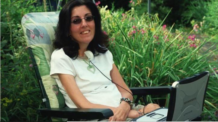 A family photo of Sarah, sitting in a leafy garden on an upright sun lounger. She is smiling and has her eye gaze technology screen on her lap. She wears a white short-sleeved top, sunglasses and a bracelet on her wrist.  