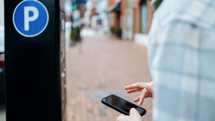 A motorist uses a mobile phone to pay for parking with an app. They are standing in front of a parking machine and looking down at the phone.A shopping street is visible in the background.