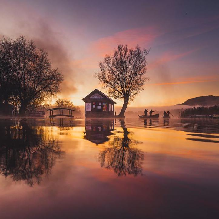 Trees and fishermen in a boat are reflected in the water. The pink and yellow sky is also reflected in the water and a little house and small bridge are to the left of the boat.