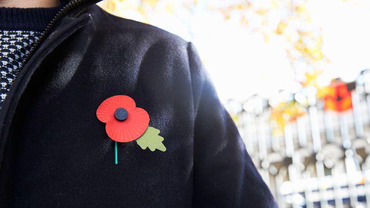 Close Up Of Man Wearing Remembrance Day Poppy. The background is blurred. He is wearing a black coat. 