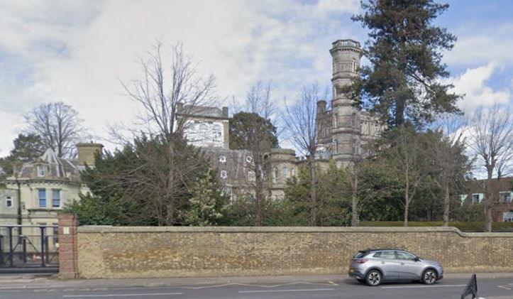 Part of Immanuel College in Bushey, seen from the road opposite. A series of quite grand historical buildings sits behind a long brick wall, including one grey building with a five storey tower. There are trees behind the wall and in the distance, and a road in the foreground.