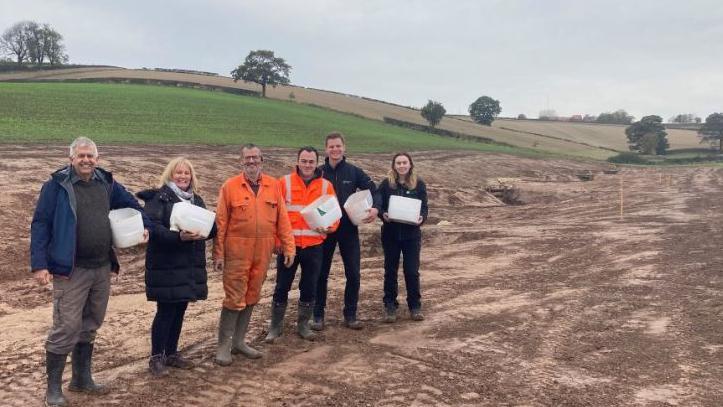 Peter Wiles and Amanda Mellor from the flood action group; John Charles-Jones, Landowner, Joshua Wells from the county council and Benjamin Doughty and Amy Facer from the Environment Agency standing in a line in a field where earth has been exposed during work on the small waterway in the background