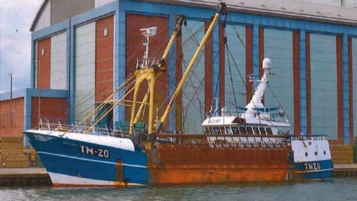 A blue and white fishing vessel with large panels of rust docked in a harbor
