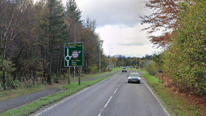 A82 leading up to the Ballachulish roundabout. There are trees at either side of the road and a sign saying a roundabout is coming up. There are two cars on the road.