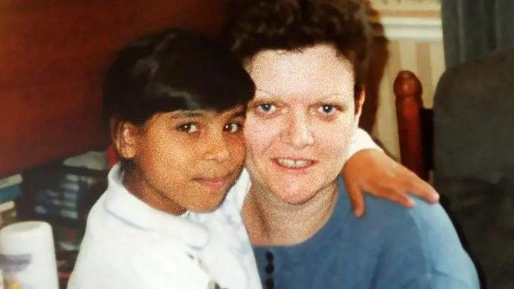 A woman and young child with their faces pressed together. The child has their arm around the woman's neck. The woman has short brown hair and is wearing a blue shirt