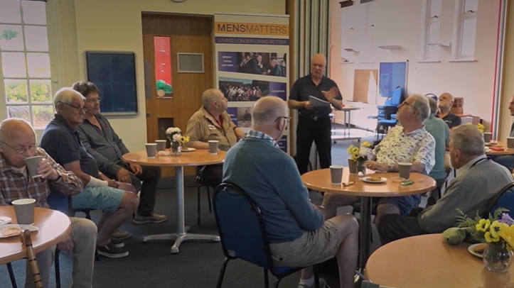 A group of men aged between 60 and 70, are sitting at several tables drinking tea, eating biscuits and taking part in a quiz.