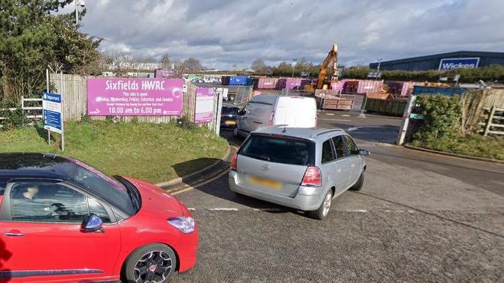 Entrance to Sixfields recycling centre showing a purple Sixfields HWRC sign on a fence to the left, and a queue of cars and vans heading through the gate.  There is a yellow digger in the background and a row of shipping containers.