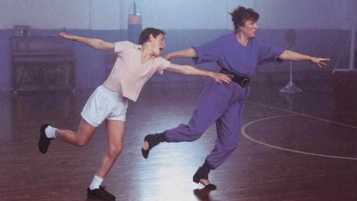 A scene from the film Billy Elliot shows Jamie Bell as Billy, dancing with Julie Walter, who plays his dance teacher Mrs Wilkinson. They are in a school gym. Billy is wearing a peach-coloured t-shirt, white shorts and socks, and black shoes. He has short dark hair. Mrs Wilkinson is wearing a purple jumpsuit with a black belt tied around her waist. She is wearing black shoes and ankle warmers. Her hair is short and red.