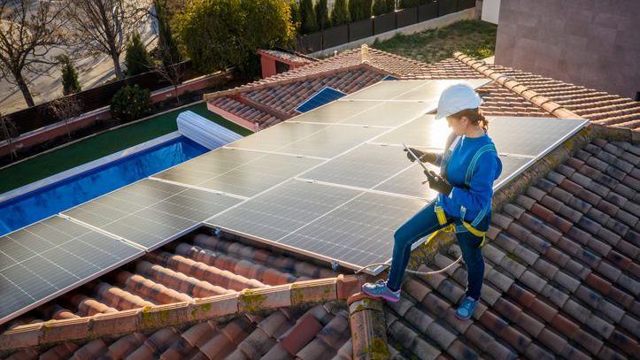 A woman works on the maintenance of a solar photovoltaic panel installed on domestic home rooftop. She is standing on the roof and looking at a tablet and it is sunset.